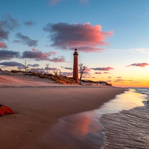 May include: A red brick lighthouse stands tall on a sandy beach with a calm ocean in the background. The sky is a vibrant mix of pink, orange, and blue, creating a beautiful sunset.