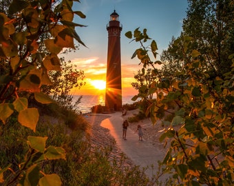 May include: A scenic sunset view featuring a tall, brick lighthouse with a bright light at the top. The sun sets over the water, casting a golden glow. Two people walk along a path towards the lighthouse, framed by trees and foliage.
