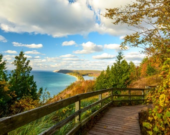 May include: A wooden walkway with a railing overlooks a blue lake and a green forested shoreline. The sky is blue with white clouds. The walkway is surrounded by trees with fall foliage.