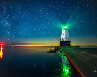 May include: A white lighthouse with a green light shining out to sea, under a starry night sky with the Milky Way visible. The lighthouse is on a pier extending into the water.