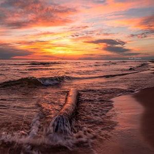 May include: A long, weathered log lies partially submerged in the sand on a beach. The waves are gently lapping at the shore, creating a soft, foamy texture. The sky is a vibrant orange and pink, with clouds reflecting the warm sunset colors.