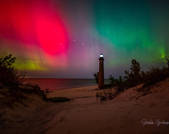 May include: A lighthouse stands tall on a sandy beach under a vibrant green and red aurora borealis. The sky is filled with stars.