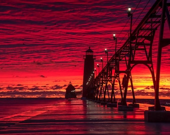 May include: A silhouette of a lighthouse and pier with a red and orange sunset in the background. The pier has lights along the side and the water is reflecting the colors of the sunset.