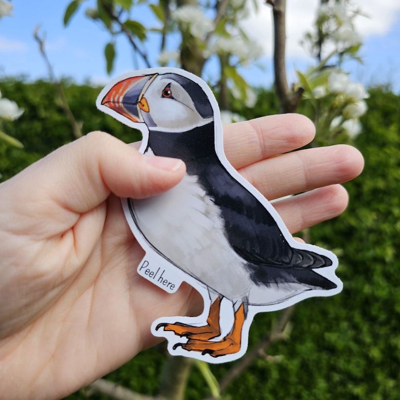 May include: A white and black puffin sticker with an orange beak. The sticker has a white background and the text "Peel here" is printed on the bottom.