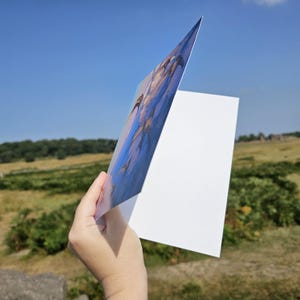 Puede incluir: Una tarjeta de felicitaci&oacute;n se sostiene contra un fondo de cielo azul y paisaje verde. La tarjeta presenta una pintura con tonos azules y dorados. El lado derecho de la tarjeta es blanco.