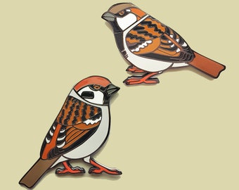 May include: Two enamel pins of sparrows, one brown and orange with black stripes, the other white and orange with black stripes. Both have black eyes and red beaks.