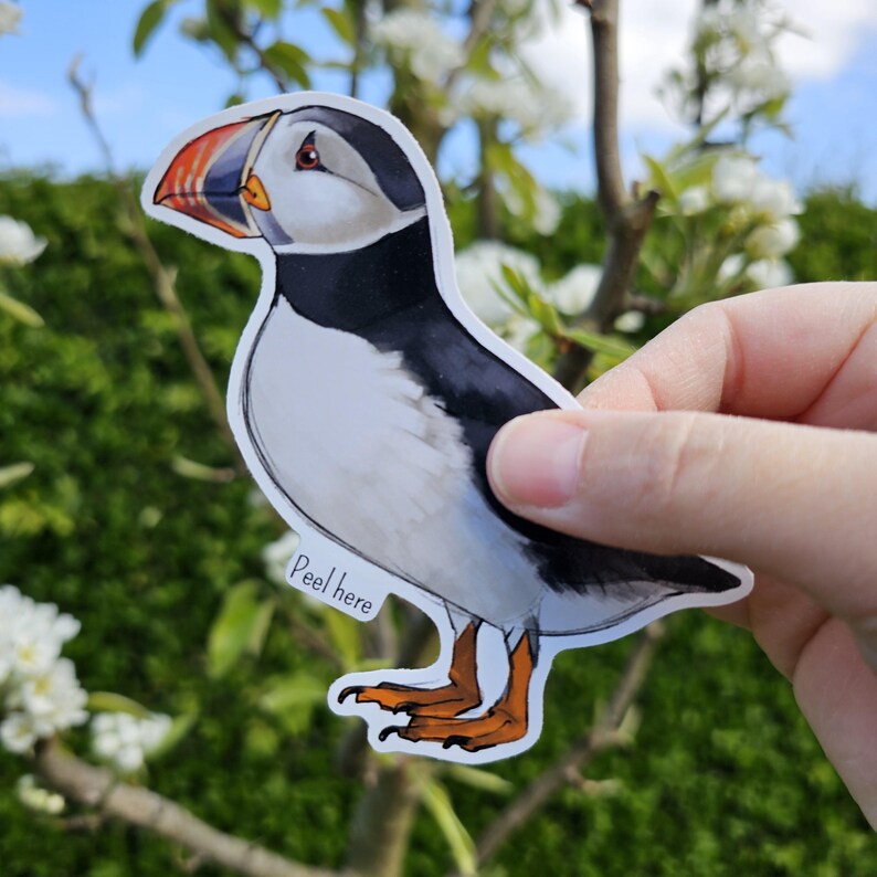 May include: A colorful sticker of a puffin with a black and white head and a white body with orange feet. The sticker has the words "Peel here" printed on it.