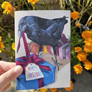 May include: A greeting card featuring a detailed illustration of a black raven holding a dark pink ribbon in its beak. The bird stands atop a blue gift box with a large bow and a white tag that reads "HAPPY BIRTHDAY" in colourful letters.
