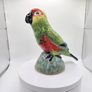 Könnte beinhalten: Keramik-Papageienfigur in Rot, Grün und Schwarz. Der Vogel sitzt auf einem strukturierten, blau-grünen Sockel. Der Kopf des Papageis ist rot und gelb, mit einem schwarzen Schnabel und Auge.
