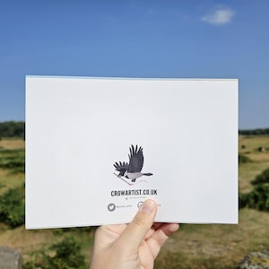Puede incluir: Tarjeta blanca con una ilustraci&oacute;n de un cuervo negro y gris sosteniendo una ramita. La tarjeta muestra el texto "CROWARTIST.CO.UK" y detalles de redes sociales. El fondo muestra un cielo azul y un paisaje verde.