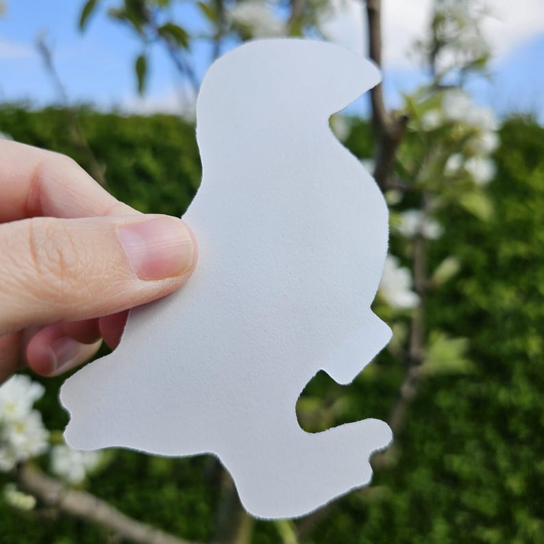 May include: White paper cut-out of a bird in a profile view. The bird is facing to the right. The cut-out is held in a hand against a background of green foliage and a blue sky.