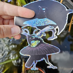 Könnte beinhalten: Zwei holografische Aufkleber mit einem stilisierten Paradiesvogel-Design. Die Vögel sind hauptsächlich schwarz mit schillernden Akzenten, die einen schimmernden Effekt erzeugen. Die Aufkleber werden vor einem verschwommenen Hintergrund aus Grün gehalten.