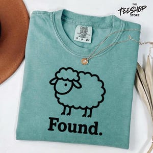 Può includere: Una maglietta Comfort Colors verde acqua con un disegno a contorno nero di una pecora e la parola "Found". La maglietta ha un girocollo e maniche corte. Una collana d'argento con un piccolo ciondolo è drappeggiata sulla maglietta.