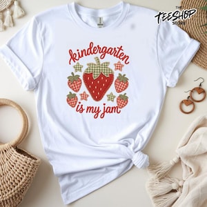 Peut inclure: Un t-shirt blanc à manches courtes avec un motif central composé de cinq fraises rouges à feuilles en vichy vert et de petites étoiles. Le texte "kindergarten is my jam" est imprimé en écriture cursive rouge. Col rond et manches courtes.