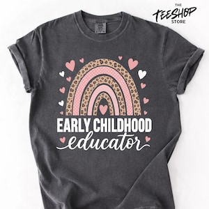 Peut inclure: T-shirt gris foncé avec un motif arc-en-ciel composé d'arches roses et léopard, entouré de cœurs roses et blancs. Le texte "EARLY CHILDHOOD educator" est imprimé en blanc. Le logo TeeShop est en haut à droite.
