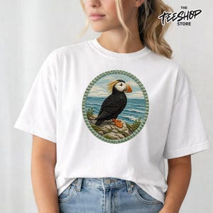 May include: A white short-sleeve t-shirt featuring a circular graphic print of a black puffin with an orange beak standing on a rocky cliff overlooking a blue ocean. The graphic is framed by a textured green border. The text THE TEESHOP STORE is visible.