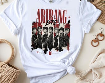 May include: A white short-sleeved t-shirt featuring a graphic print of the music group BTS. The design includes the text ARIRANG in large red letters above black and white portraits of the members, set against vertical red and white stripes.