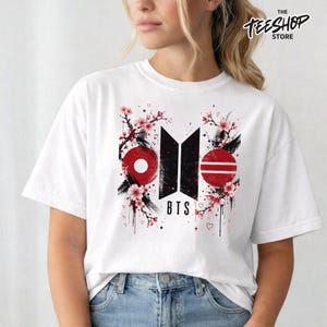 May include: A white short-sleeved t-shirt featuring a central graphic design with the black BTS logo, red circular accents, and pink cherry blossom branches. The design includes small red hearts and the text BTS printed in a bold black font.