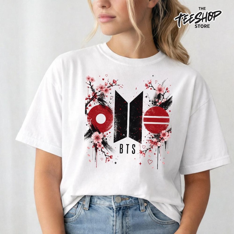 May include: A white short-sleeved t-shirt featuring a central graphic design with the black BTS logo, red circular accents, and pink cherry blossom branches. The design includes small red hearts and the text BTS printed in a bold black font.