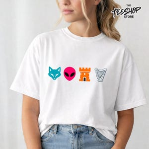 Peut inclure: T-shirt blanc avec quatre icônes graphiques colorées : une tête de renard turquoise, une tête d'extraterrestre rose, un château orange et une harpe grise. Le texte "THE TEESHOP STORE" est imprimé en noir en haut à droite.