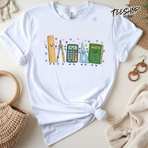 Peut inclure: Un t-shirt blanc à manches courtes orné d'un motif coloré représentant des fournitures scolaires de dessin animé, dont une règle, un compas, une calculatrice, une équerre et un livre vert marqué MATH, tous souriants, entourés d'étoiles.