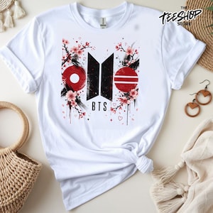 May include: A white short-sleeved t-shirt featuring a graphic print with the black BTS logo, red circular accents, and pink cherry blossom branches. The design includes black ink-like splatters, small hearts, and the text The Teeshop Store.