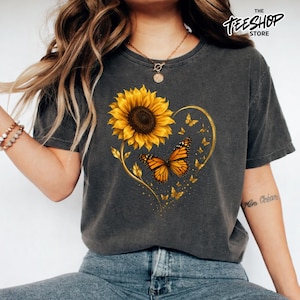 Peut inclure: Un t-shirt gris anthracite orné d'un grand tournesol jaune et d'un papillon monarque orange disposés en forme de cœur. Le motif comprend des silhouettes de papillons dorés plus petits et le texte THE TEESHOP STORE en haut à droite.
