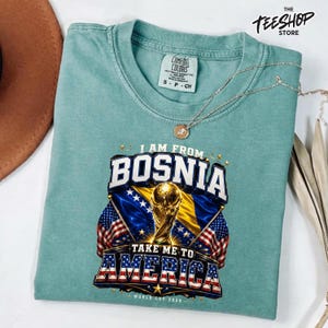 Peut inclure: Un t-shirt à col rond vert mousse avec une impression graphique portant le texte I AM FROM BOSNIA TAKE ME TO AMERICA WORLD CUP 2026. Le motif inclut un trophée doré, le drapeau bosnien et le drapeau américain. Un collier argenté est posé dessus.