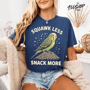 Peut inclure: T-shirt bleu marine avec un dessin d'une perruche verte et jaune sur des graines, avec le texte "SQUAWK LESS SNACK MORE" en jaune. Le logo The TeeShop Store est visible.