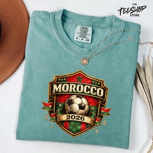 Op de afbeelding: Een saliegroen t-shirt met een afbeelding van een voetbal in het midden van een schildembleem. Het schild toont de tekst MOROCCO en 2026, geaccentueerd met rode en groene strepen, gouden sterren en decoratieve linten. Een zilveren ketting ligt op het shirt.
