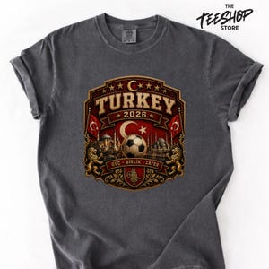 Peut inclure: Un t-shirt gris anthracite avec une impression graphique portant le texte TURKEY 2026, un ballon de football, un croissant de lune et une étoile, des silhouettes architecturales, deux lions et les mots GÜÇ, BIRLIK, ZAFER dans un emblème.