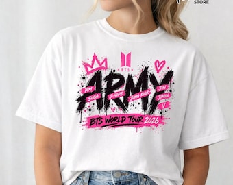 May include: A white short-sleeve t-shirt featuring a black brushstroke graphic that reads ARMY with pink accents, a crown, and a heart. The text includes the names RM, SUGA, J-HOPE, JUNG KOOK, JIMIN, V, and the text BTS WORLD TOUR 2026.