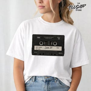 Peut inclure: Un t-shirt blanc à manches courtes avec un motif de cassette audio noire TDK SA-X 60. L'étiquette de la cassette porte le texte manuscrit DJ-Screw, June 27 et Fool, ainsi que le numéro 87-1092 imprimé sur le côté inférieur gauche.