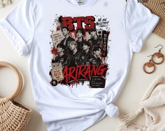 May include: A white short-sleeved t-shirt featuring a graphic print of the music group BTS. The design includes the text BTS, ARIRANG, and WE ARE BULLETPROOF, along with images of the seven members, red paint splatter effects, and various ticket-style labels.