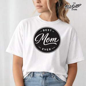 Peut inclure: T-shirt blanc avec un motif circulaire noir portant l'inscription "BEST Mom EVER" en cursive. Le t-shirt est porté avec un jean bleu.