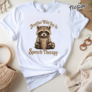 Peut inclure: Un t-shirt blanc à manches courtes avec une illustration d'un raton laveur brun tenant un gobelet en plastique transparent avec une paille. Le texte au-dessus du raton laveur indique Another Wild Day In et Speech Therapy en dessous en brun.