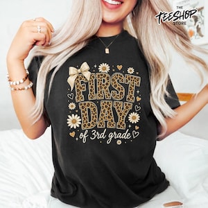Peut inclure: Un t-shirt noir à manches courtes avec une impression graphique portant le texte "FIRST DAY of 3rd grade" dans un motif léopard marron. Le design comprend un grand nœud beige, de petites marguerites blanches et des cœurs.
