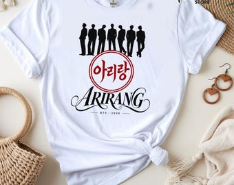 May include: A white short-sleeved t-shirt featuring a black silhouette graphic of seven figures standing in a row. Below the figures is a red circular emblem with Korean text, the word Arirang in black script, and the text BTS - 2026.