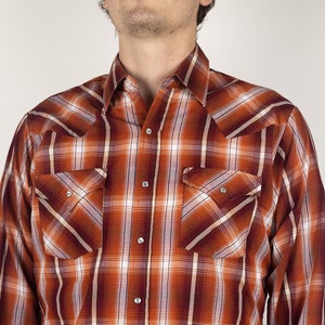 Puede incluir: Una camisa de cuadros de manga larga con cuello abotonado. La camisa es naranja, blanca y marrón con un patrón a cuadros.