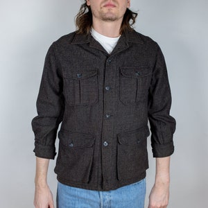 Può includere: Una camicia abbottonata con motivo marrone e nero con più tasche e colletto. La camicia è indossata sopra una t-shirt bianca e jeans blu.