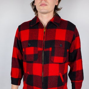 Puede incluir: Una camisa de franela a cuadros rojos y negros con cierre de cremallera. La camisa tiene dos bolsillos de solapa en el pecho y mangas largas.