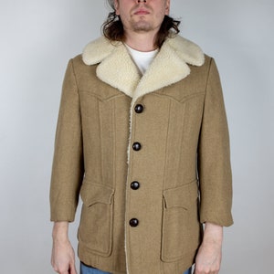 Op de afbeelding: Een bruine corduroy jas met een shearling kraag en bruine knopen. De jas heeft twee grote zakken aan de voorkant en een enkele zak op de linker borst. Het model draagt ​​een blauwe jeans en een groene baseballpet met de tekst "Miller High Life" op de voorkant.