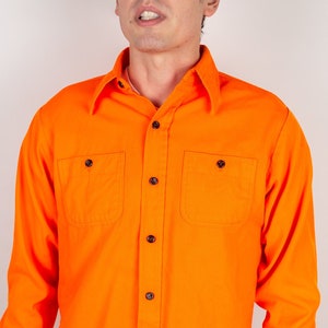 Peut inclure: Une chemise à manches longues de couleur orange vif avec un devant boutonné et deux poches poitrine. La chemise a un style décontracté, de type vêtement de travail.