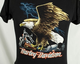 May include: Black t-shirt with a graphic of a bald eagle with outstretched wings, lightning bolts, and a Harley-Davidson motorcycle. The text "Harley-Davidson" is printed in orange below the graphic.