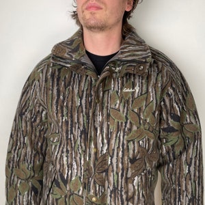 May include: A camouflage-patterned jacket with a zipper and snap closure. The jacket has a high collar and is made of a soft, fuzzy material. The brand name "Cabela's" is visible on the left chest.