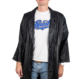 Puede incluir: Una larga gabardina de cuero negro con cierre de botones. La gabardina tiene cuello y bolsillos. La persona que lleva la gabardina también lleva una camiseta blanca con el texto "Saint ST. PAUL" en azul.