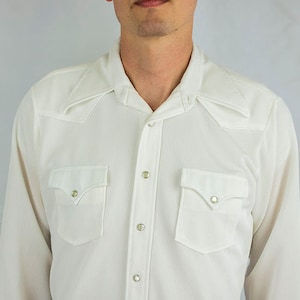 May include: A long-sleeved white button-down shirt featuring a textured fabric, a pointed collar, two chest pockets with flaps, and snap closures. The shirt has wide cuffs and a straight hemline, presenting a classic western-inspired design.