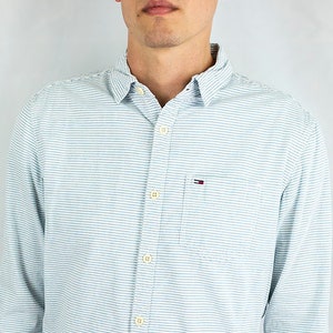 May include: A light blue striped long-sleeve button-down shirt with a small logo on the left chest pocket. The shirt is tucked into blue denim shorts.