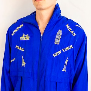 May include: A blue windbreaker jacket with a zipper closure and a drawstring at the waist. The jacket features embroidered images of famous landmarks from around the world, including the Eiffel Tower, the Statue of Liberty, Big Ben, and the Leaning Tower of Pisa. The text "LONDON", "PARIS", "NEW YORK", and "MILAN" is also embroidered on the jacket.