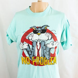 May include: A light blue t-shirt featuring a cartoon image of Bugs Bunny wearing sunglasses and a tie. The image is surrounded by a red circle with a diagonal line through it. The text "NO PROBLEM" is printed below the image.